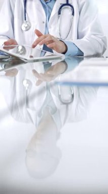 Mavi bluzlu, steteskoplu ve beyaz önlüklü tanınmayan kadın doktor modern sağlık ortamında dijital tablet kullanıyor, hasta verileriyle ve dikey görüşüyle etkileşimde. Tıp ve Bilim.