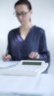Hesap makinesi ve muhasebe kağıtları cam masanın üstünde. Hesap makinesi kullanan ve arka planda not alan bir muhasebeci. Ticarette denetim ve vergiler,