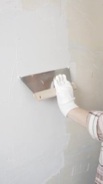 Eldiven giyen kimliği belirsiz kadın inşaat işçisi duvara sıva uygulamak için macun bıçağı kullanıyor. Ev yenileme projesi sırasında yüzeyi düzleştiriyor.