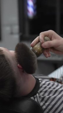 Profesyonel berber müşterinin yüzünü fırçayla süpürüyor, müşterilerin sakallarını geleneksel bir berber dükkanında tımarlanmaya hazırlıyor..