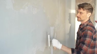 Eldiven giyen erkek inşaat işçisi, macun bıçağıyla ustalıkla duvara alçı yapıştırıyor, bir ev yenileme projesiyle meşgul, iç mekanı hassasiyetle büyütüyor.