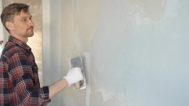 Eldiven giyen erkek inşaat işçisi, bir el aleti kullanarak duvara alçı yapıştırıyor, ev yenileme veya inşaat projesine katkıda bulunuyor