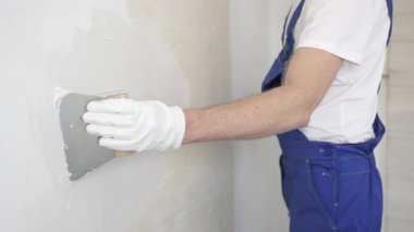 Bilinmeyen inşaat işçisi, beyaz duvar boyuyor, mavi tulum giyiyor ve apartman yenileme projesi sırasında koruyucu beyaz eldivenler giyiyor..