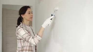 Koruyucu eldivenler giyen bir kadın, beyaz duvarı mavi bigudiyle boyuyor, iç mekanı güncelliyor ve yenileme projesinde odayı temizliyor, modern görünüme sahip..