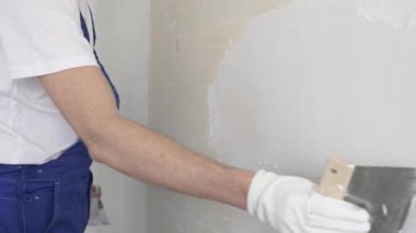 Bilinmeyen inşaat işçisi, beyaz duvar boyuyor, mavi tulum giyiyor ve apartman yenileme projesi sırasında koruyucu beyaz eldivenler giyiyor..