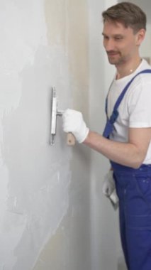 Profesyonel inşaat işçisi mavi tulum ve beyaz eldiven giyerek, yenilenmiş, dikey görünümlü bir apartman dairesine beyaz bir duvar örmek için mala kullanıyor.