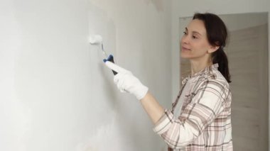 Koruyucu eldivenler giyen bir kadın, beyaz duvarı mavi bigudiyle boyuyor, iç mekanı güncelliyor ve yenileme projesinde odayı temizliyor, modern görünüme sahip..