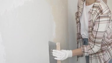 Eldiven giyen kimliği belirsiz kadın inşaat işçisi duvara sıva uygulamak için macun bıçağı kullanıyor. Ev yenileme projesi sırasında yüzeyi düzleştiriyor.