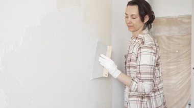 Kadın duvara macun bıçağıyla alçı yapıştırıyor, koruma için eldiven takıyor ve tadilat sırasında bile pürüzsüz ve sorunsuz bir şekilde bitmesini sağlıyor.