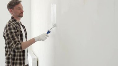 Profesyonel ressam beyaz duvarı silindirle düzleştiriyor, ev dekorasyonu sırasında koruyucu eldiven takıyor, iç mekanı yeni boyama tekniğiyle değiştiriyor.