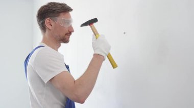 Güvenlik gözlüğü ve çalışma eldiveni takan inşaat işçisi, bir ev yenileme veya inşaat projesi sırasında ustalıkla duvara çivi çakıyor, hassasiyet ve güvenliğe odaklanıyor.