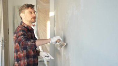 Eldiven giyen erkek inşaat işçisi, bir el aleti kullanarak duvara alçı yapıştırıyor, ev yenileme veya inşaat projesine katkıda bulunuyor