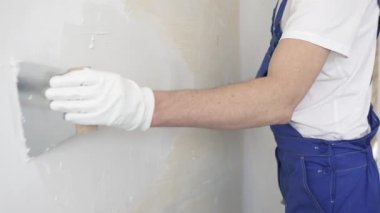 Bilinmeyen inşaat işçisi, beyaz duvar boyuyor, mavi tulum giyiyor ve apartman yenileme projesi sırasında koruyucu beyaz eldivenler giyiyor..