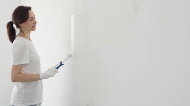 Beyaz eldiven giyen mutlu bir kadın boya silindirli beyaz bir duvar boyuyor. Ev yenileme sürecinin tadını çıkarıyor ve yeni bir görünüm yaratıyor.