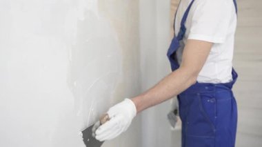 Bilinmeyen inşaat işçisi, beyaz duvar boyuyor, mavi tulum giyiyor ve apartman yenileme projesi sırasında koruyucu beyaz eldivenler giyiyor..