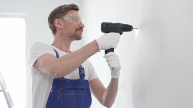 Profesyonel tamirci mavi üniformalı, güvenlik gözlüğü ve eldiven takıyor, tadilat ve montaj sırasında beyaz duvarda delik açmak için kablosuz matkap kullanıyor..