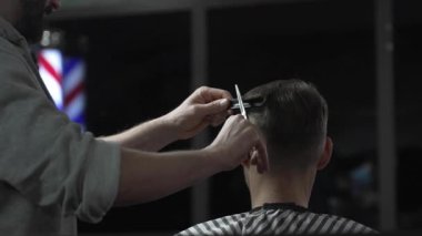 Profesyonel berber erkek müşterinin saçını şık bir berber dükkanında makas ve tarak kullanarak ustaca kesiyor ve arka planda belirgin bir şekilde berber direği sergiliyor.