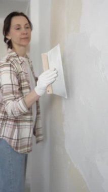 Kadın duvara macun bıçağıyla alçı yapıştırıyor, koruma için eldiven takıyor ve ev tadilatı, dikey görünüm süresince pürüzsüz ve pürüzsüz bir şekilde bitmesini sağlıyor.