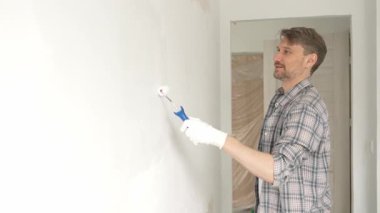 Orta yaşlı bir adam, apartman duvarında küçük bir silindirle beyaz boya yuvarlıyor. Geniş kapsamlı ev yenileme çalışmaları sırasında sürekli darbeler alarak iç mekanı değiştiriyor..