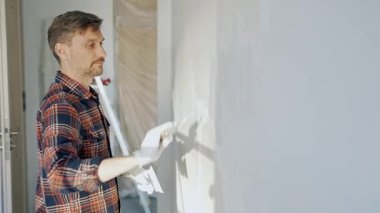 Eldiven giyen erkek inşaat işçisi, bir el aleti kullanarak duvara alçı yapıştırıyor, ev yenileme veya inşaat projesine katkıda bulunuyor