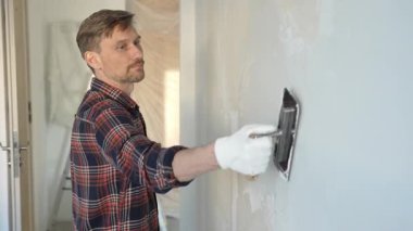 Eldiven giyen erkek inşaat işçisi, bir el aleti kullanarak duvara alçı yapıştırıyor, ev yenileme veya inşaat projesine katkıda bulunuyor