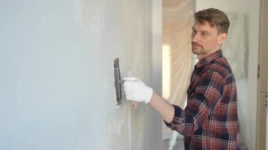 Eldiven giyen erkek inşaat işçisi, macun bıçağıyla ustalıkla duvara alçı yapıştırıyor, bir ev yenileme projesiyle meşgul, iç mekanı hassasiyetle büyütüyor.