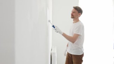 Beyaz eldiven ve tişört giyen orta yaşlı bir adam, boya silindirli beyaz bir duvar boyuyor, evini yeniliyor ve yaşam alanını dönüştürüyor..