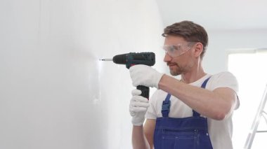 Mavi tulumlu, güvenlik gözlüklü ve iş eldivenli bir inşaat işçisi kablosuz tornavida kullanarak duvara vida sokuyor.