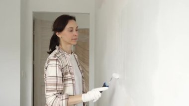 Koruyucu eldivenler giyen bir kadın, beyaz duvarı mavi bigudiyle boyuyor, iç mekanı güncelliyor ve yenileme projesinde odayı temizliyor, modern görünüme sahip..