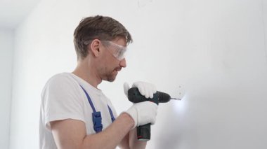 Güvenlik gözlüğü ve eldiven takan profesyonel tamirat ve montaj sırasında beyaz duvarda delik açmak için kablosuz matkap kullanıyor.