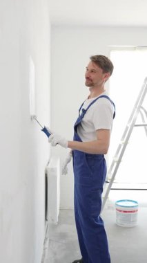 Beyaz eldivenler ve mavi tulumlar giyen profesyonel boyacı boş bir odada temiz beyaz bir duvar boyuyor, yenileme ve iç dekorasyona odaklanıyor.