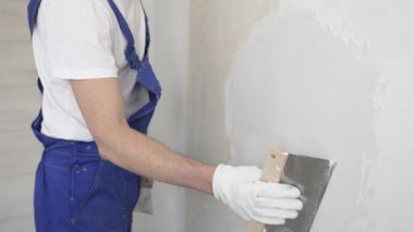 Bilinmeyen inşaat işçisi, beyaz duvar boyuyor, mavi tulum giyiyor ve apartman yenileme projesi sırasında koruyucu beyaz eldivenler giyiyor..