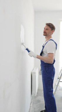 Beyaz eldivenler ve mavi tulumlar giyen profesyonel boyacı boş bir odada temiz beyaz bir duvar boyuyor, yenileme ve iç dekorasyona odaklanıyor.
