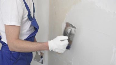 Bilinmeyen inşaat işçisi, beyaz duvar boyuyor, mavi tulum giyiyor ve apartman yenileme projesi sırasında koruyucu beyaz eldivenler giyiyor..