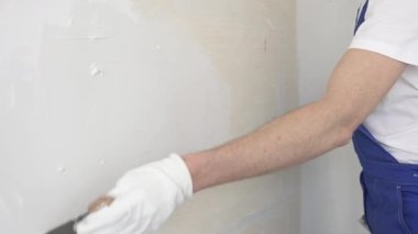 Bilinmeyen inşaat işçisi, beyaz duvar boyuyor, mavi tulum giyiyor ve apartman yenileme projesi sırasında koruyucu beyaz eldivenler giyiyor..