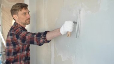 Eldiven giyen erkek inşaat işçisi, bir el aleti kullanarak duvara alçı yapıştırıyor, ev yenileme veya inşaat projesine katkıda bulunuyor