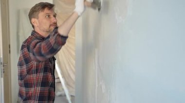 Eldiven giyen erkek inşaat işçisi, bir el aleti kullanarak duvara alçı yapıştırıyor, ev yenileme veya inşaat projesine katkıda bulunuyor