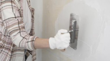Eldiven giyen kimliği belirsiz kadın inşaat işçisi duvara sıva uygulamak için macun bıçağı kullanıyor. Ev yenileme projesi sırasında yüzeyi düzleştiriyor.