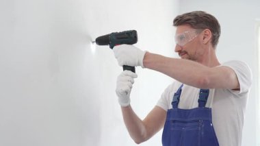 Profesyonel tamirci mavi üniformalı, güvenlik gözlüğü ve eldiven takıyor, tadilat ve montaj sırasında beyaz duvarda delik açmak için kablosuz matkap kullanıyor..