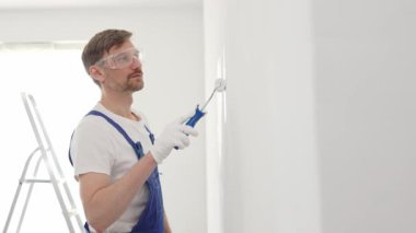Beyaz eldivenler ve mavi tulumlar giyen profesyonel boyacı boş bir odada temiz beyaz bir duvar boyuyor, yenileme ve iç dekorasyona odaklanıyor.