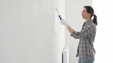 Eldiven giyen bir kadın boya silindiriyle beyaz bir duvar boyuyor, evini canlı bir ceketle değiştiriyor ve yenileme ve geliştirme sürecinin tadını çıkarıyor.