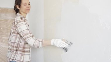 Kadın duvara macun bıçağıyla alçı yapıştırıyor, koruma için eldiven takıyor ve tadilat sırasında bile pürüzsüz ve sorunsuz bir şekilde bitmesini sağlıyor.