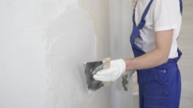 Bilinmeyen inşaat işçisi, beyaz duvar boyuyor, mavi tulum giyiyor ve apartman yenileme projesi sırasında koruyucu beyaz eldivenler giyiyor..
