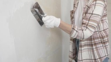 Eldiven giyen kimliği belirsiz kadın inşaat işçisi duvara sıva uygulamak için macun bıçağı kullanıyor. Ev yenileme projesi sırasında yüzeyi düzleştiriyor.