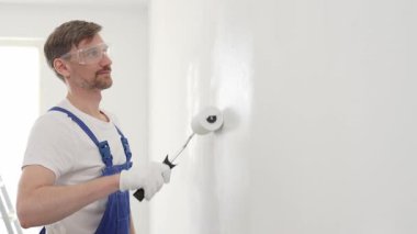 Mavi üniformalı erkek profesyonel ressam, iç dekorasyon projesi sırasında düzgün bir yüzey oluşturarak, duvarda dikkatlice beyaz boya sürüyor..
