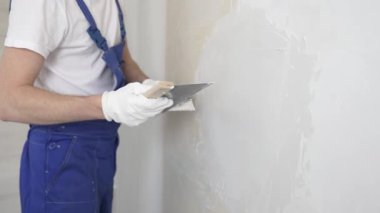 Bilinmeyen inşaat işçisi, beyaz duvar boyuyor, mavi tulum giyiyor ve apartman yenileme projesi sırasında koruyucu beyaz eldivenler giyiyor..
