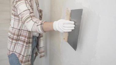 Eldiven giyen kimliği belirsiz kadın inşaat işçisi duvara sıva uygulamak için macun bıçağı kullanıyor. Ev yenileme projesi sırasında yüzeyi düzleştiriyor.