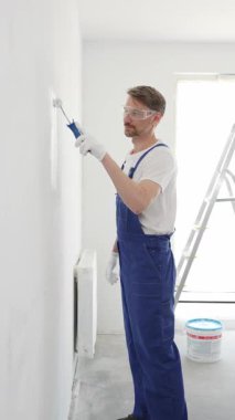 Beyaz eldivenler ve mavi tulumlar giyen profesyonel boyacı boş bir odada temiz beyaz bir duvar boyuyor, yenileme ve iç dekorasyona odaklanıyor.
