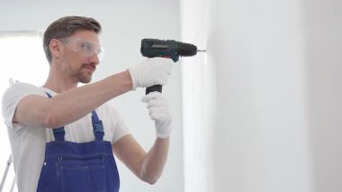 Profesyonel tamirci mavi üniformalı, güvenlik gözlüğü ve eldiven takıyor, tadilat ve montaj sırasında beyaz duvarda delik açmak için kablosuz matkap kullanıyor..