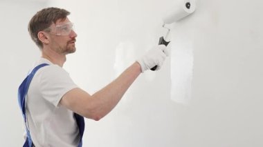 Mavi üniformalı erkek profesyonel ressam, iç dekorasyon projesi sırasında düzgün bir yüzey oluşturarak, duvarda dikkatlice beyaz boya sürüyor..